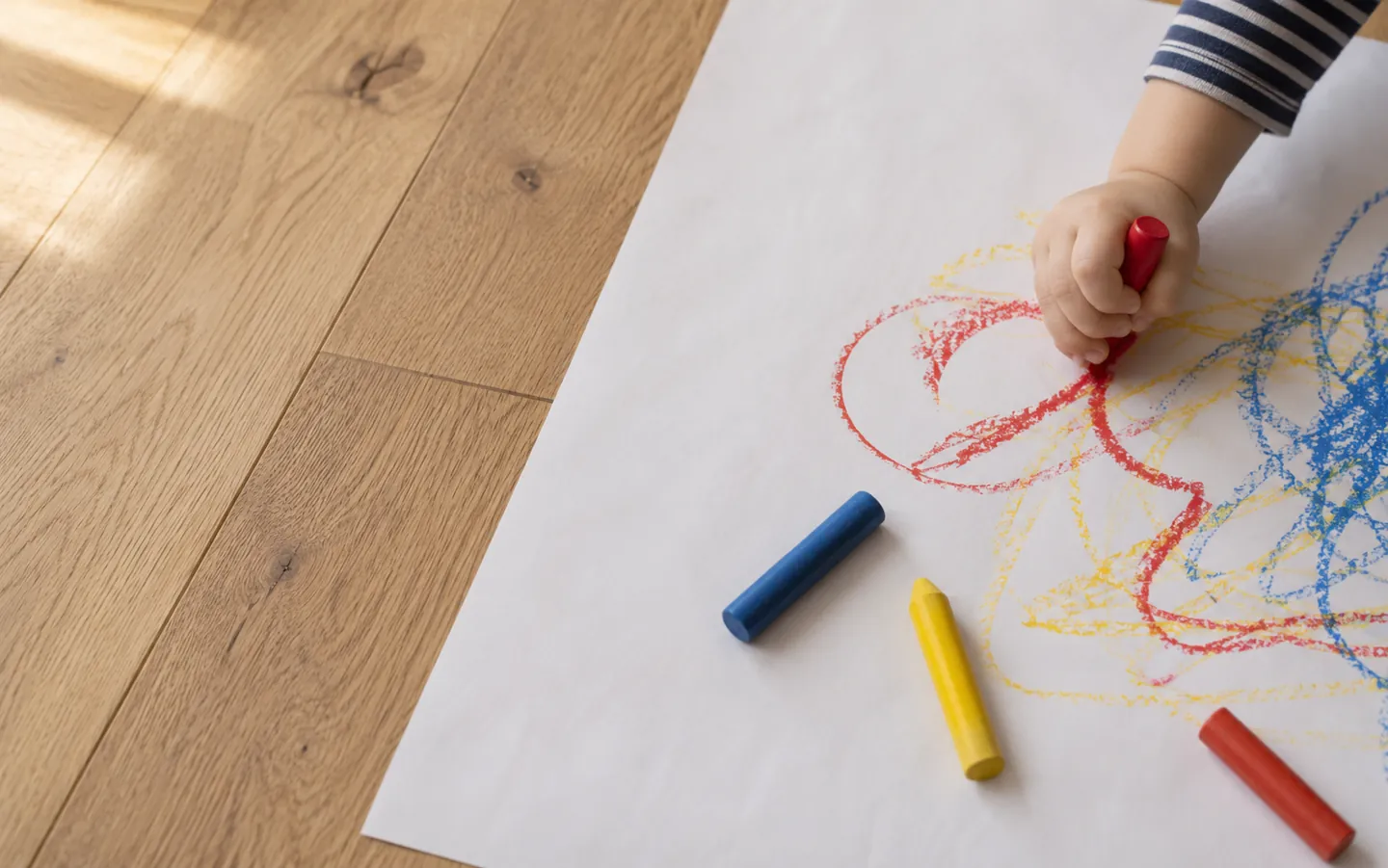 A toddler's hand gripping a chunky wax crayon and scribbling bright loops across a large sheet of paper on a wooden floor